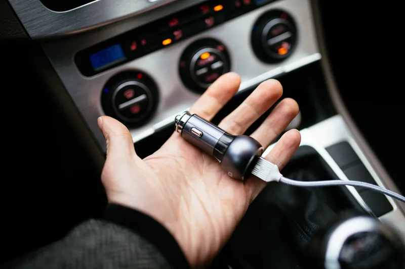 A hand holding a USB car charger in front of a 12V power outlet.